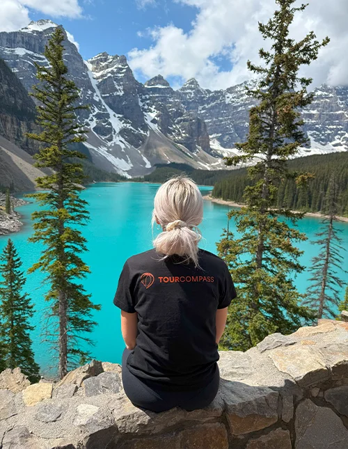 Eine Frau im TourCompass-T-Shirt blickt auf einen türkisfarbenen See und schneebedeckte Berge in Kanada