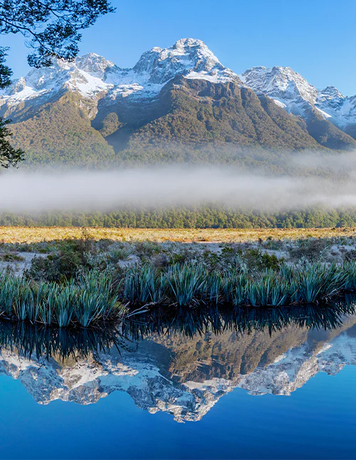 Spiegelsee, tief hängender Nebel und schneebedeckte Gipfel an einem klaren Wintertag im Milford Sound, Neuseeland